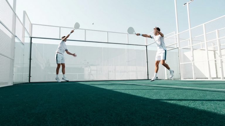 Deux joueurs de padel en tenue blanche sur un court extérieur vert. Ils échangent la balle, raquettes en main, sous un ciel clair.